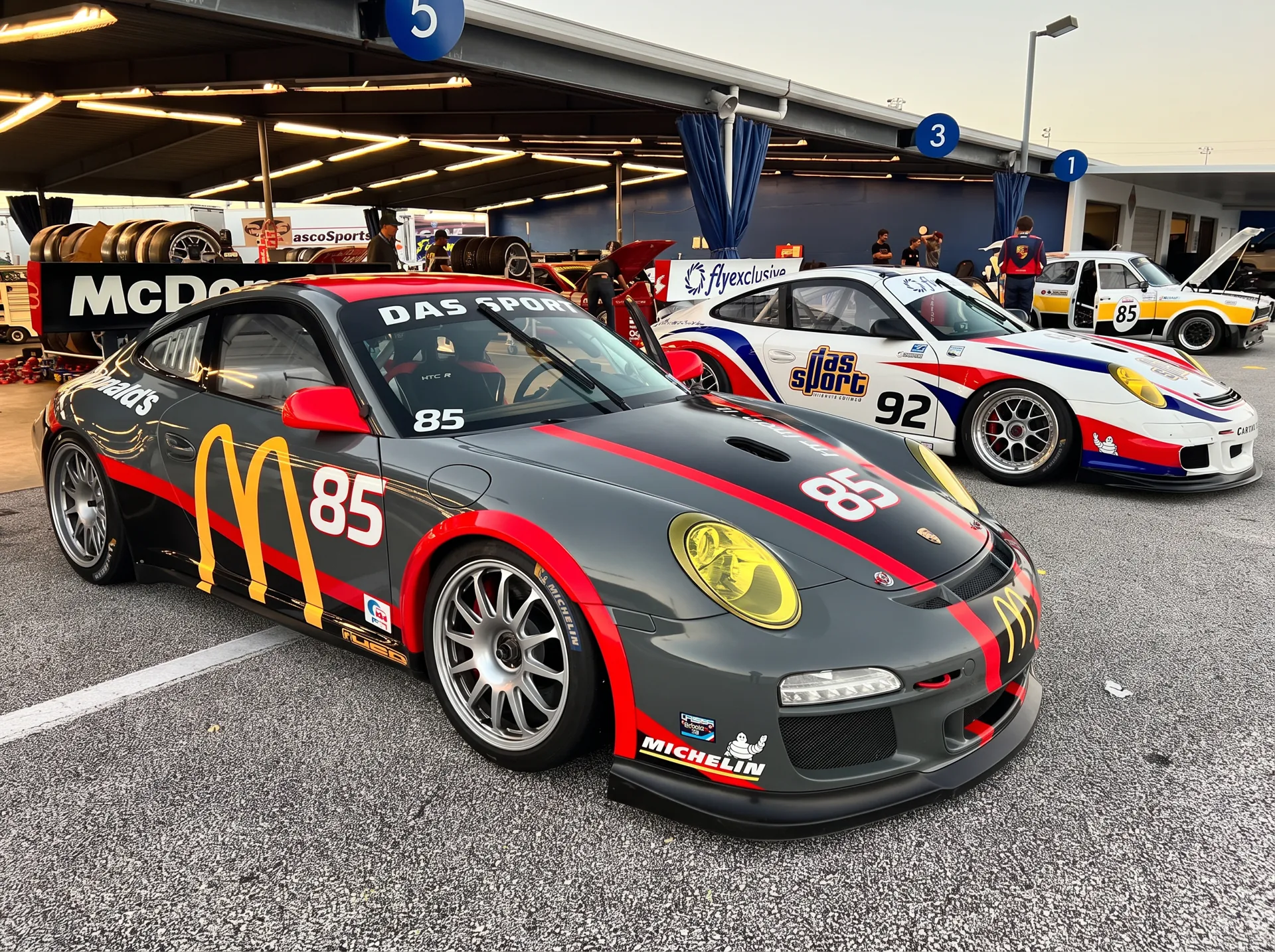 DAS Sport #85 and #92 Porsche race cars staged in the paddock garages at sunset